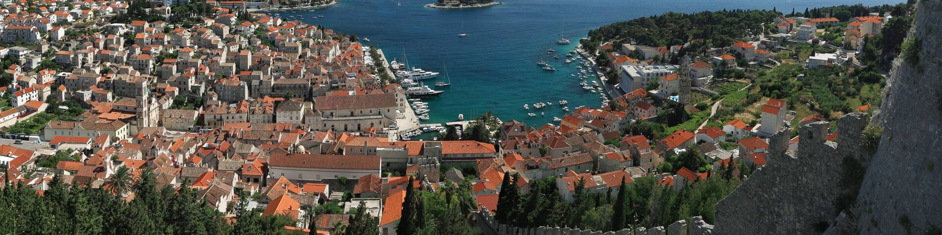 A coastal town with red-roofed buildings, a historic stone wall, and a clear blue sea with boats.
