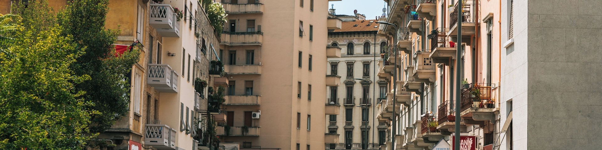 A narrow urban street lined with multi-story buildings, parked cars, and a few trees.