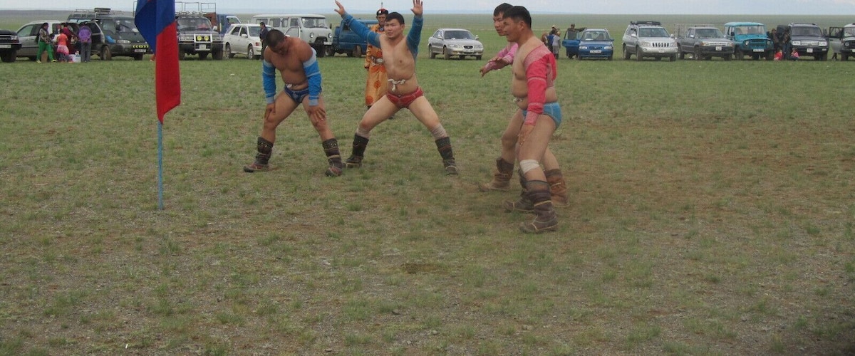 Three men in traditional attire performing a dance in an open field with vehicles and a flag in the background.