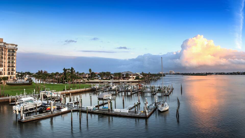 A marina with boats docked, a large building on the left, and a city skyline in the background.