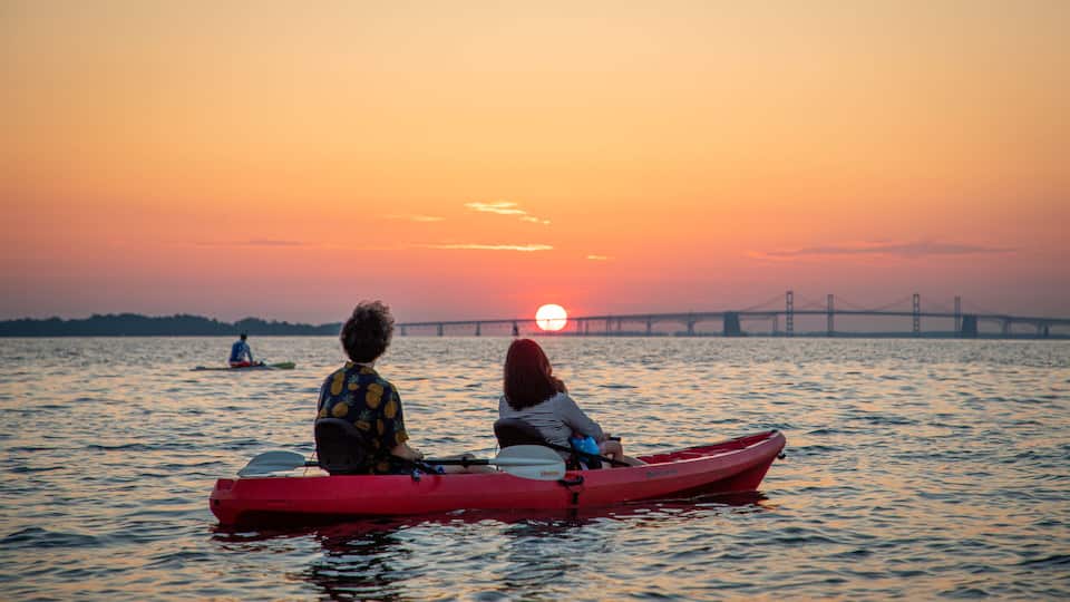 Two people in a red canoe on a body of water with a sunset and a bridge in the background.
