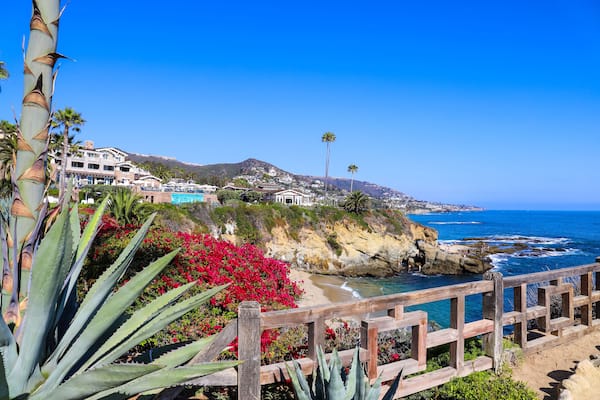 a gorgeous shot of the vast deep blue ocean water with waves rolling into the beach, homes on the cliffs with lush green palm trees and plants with blue sky at Treasure Island Beach in Laguna Beach CA