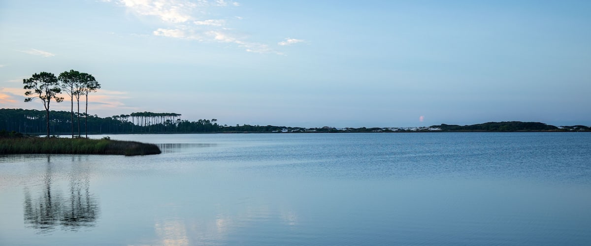 Un llac tranquil amb arbres alts que es reflecteixen a l'aigua, envoltat de turons i un cel serè.