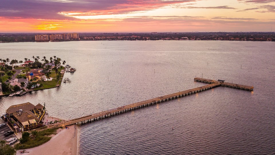 A sunset over a large body of water with a long pier extending into it, surrounded by residential buildings and a small island with a dock.