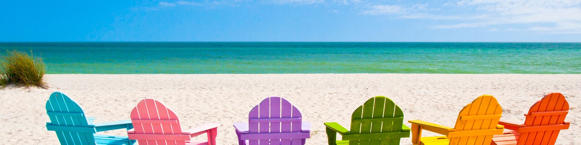 Una hilera de sillas Adirondack coloridas en una playa arenosa, con cielo azul claro y océano turquesa.