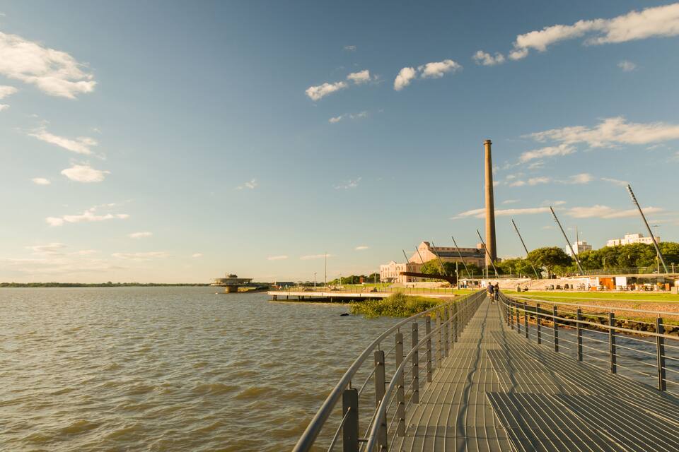 A riverside walkway with a metal railing, a tall chimney, and a bridge in the background.