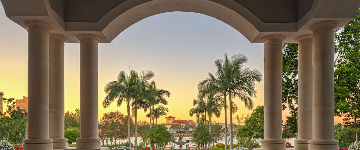 A fountain surrounded by palm trees and flowers under a large archway.