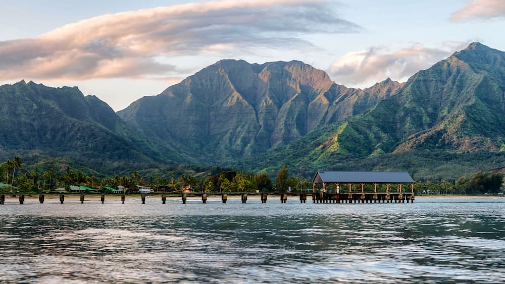 A wooden pier extending into a calm lake, with a small pavilion at the end. In the background, there are lush green mountains and a few palm trees.