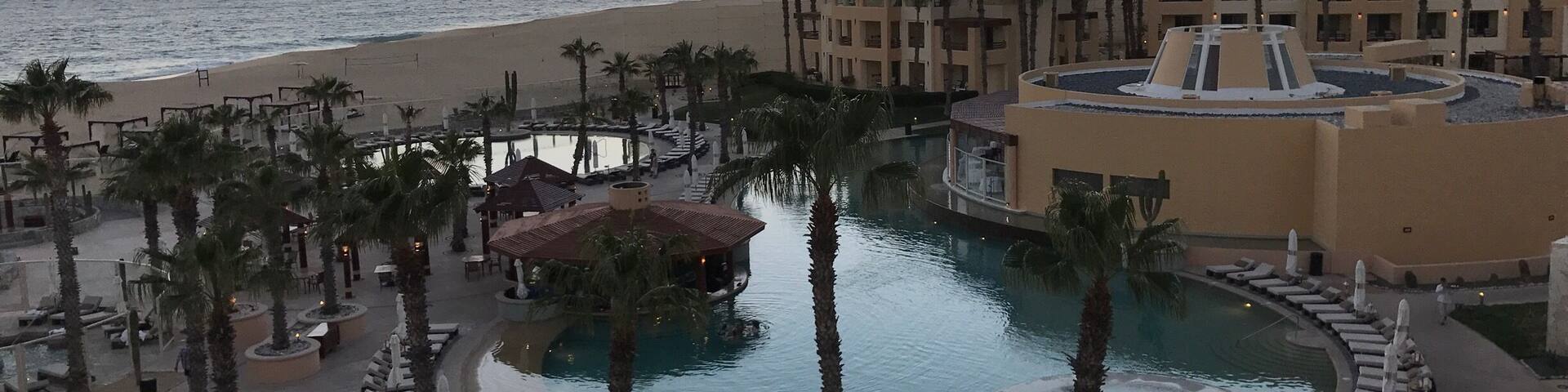 A resort with a pool, palm trees, and a beach at sunset.