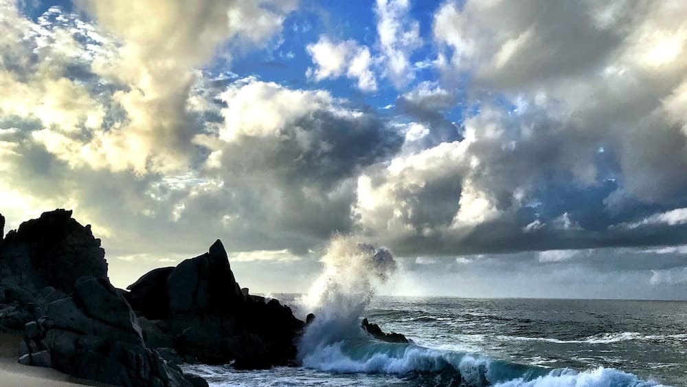 Une plage où les vagues s’écrasent contre les rochers sous un ciel nuageux.