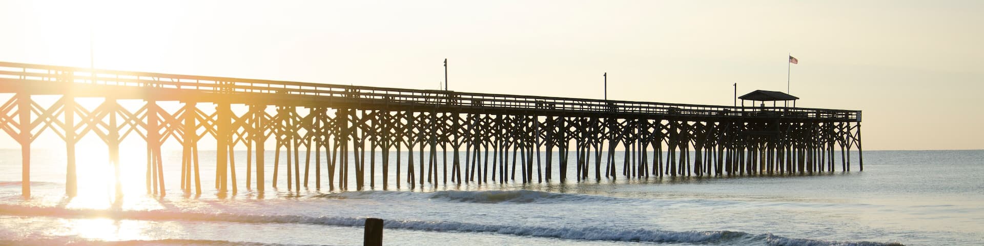 Looking east into a golden sunrise with the tall timber structure of a pier in silhouette against the bright sun, casting long shadows on the sand.
