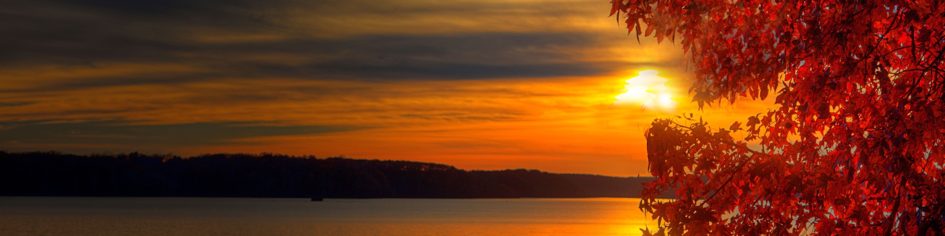 A sunset over a calm lake with red autumn leaves on the right.