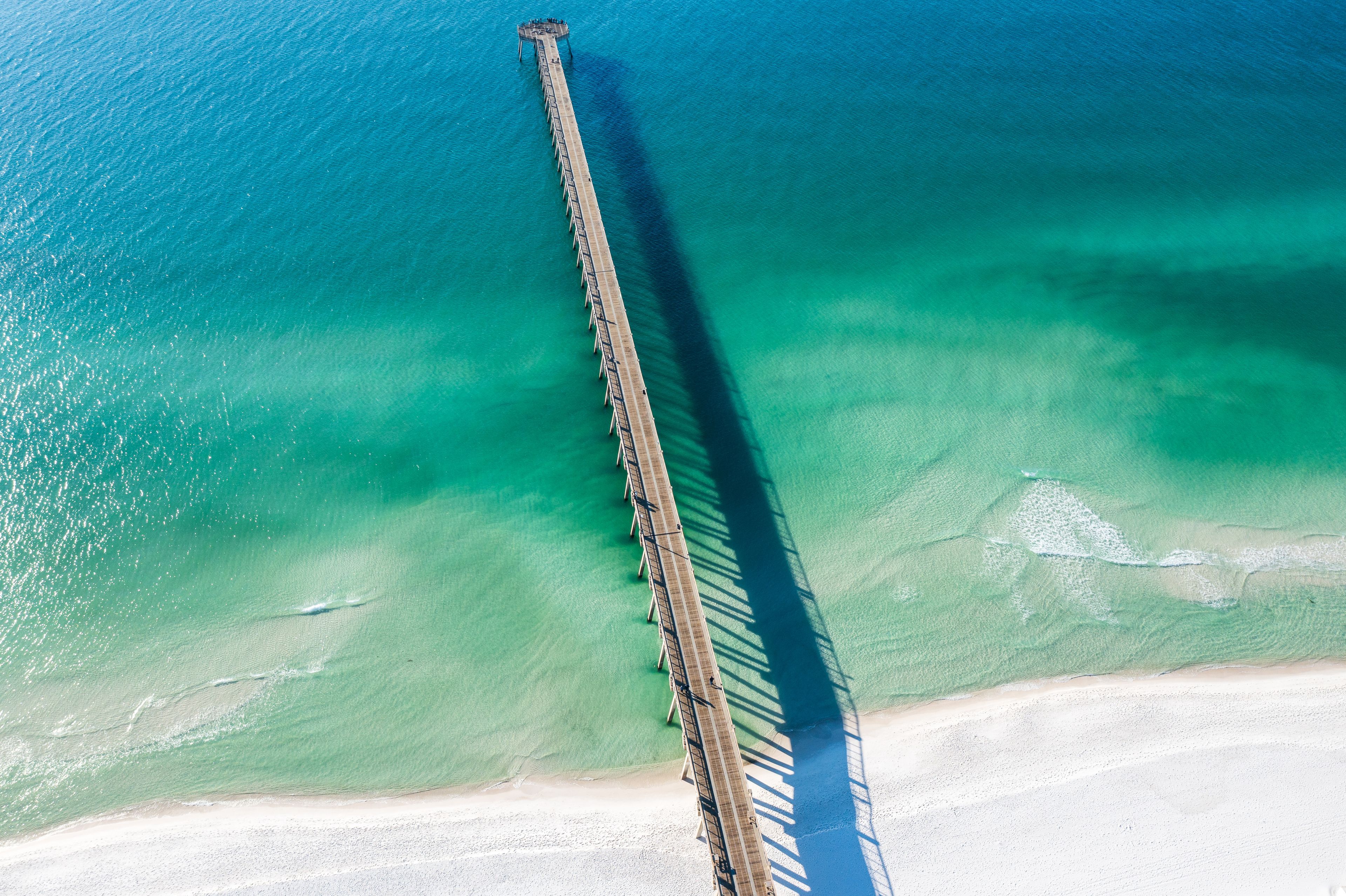 Vista aérea de un muelle largo que se extiende sobre un mar turquesa con una playa de arena al final.