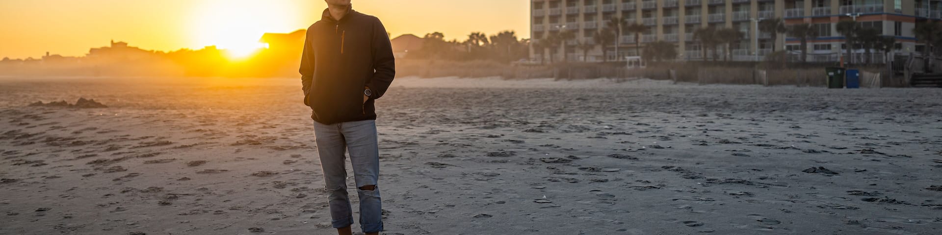 One young man silhouette standing watching colorful sunset at Myrtle Beach city by Atlantic ocean with sun behind horizon by condo apartment buildings in South Carolina resort town