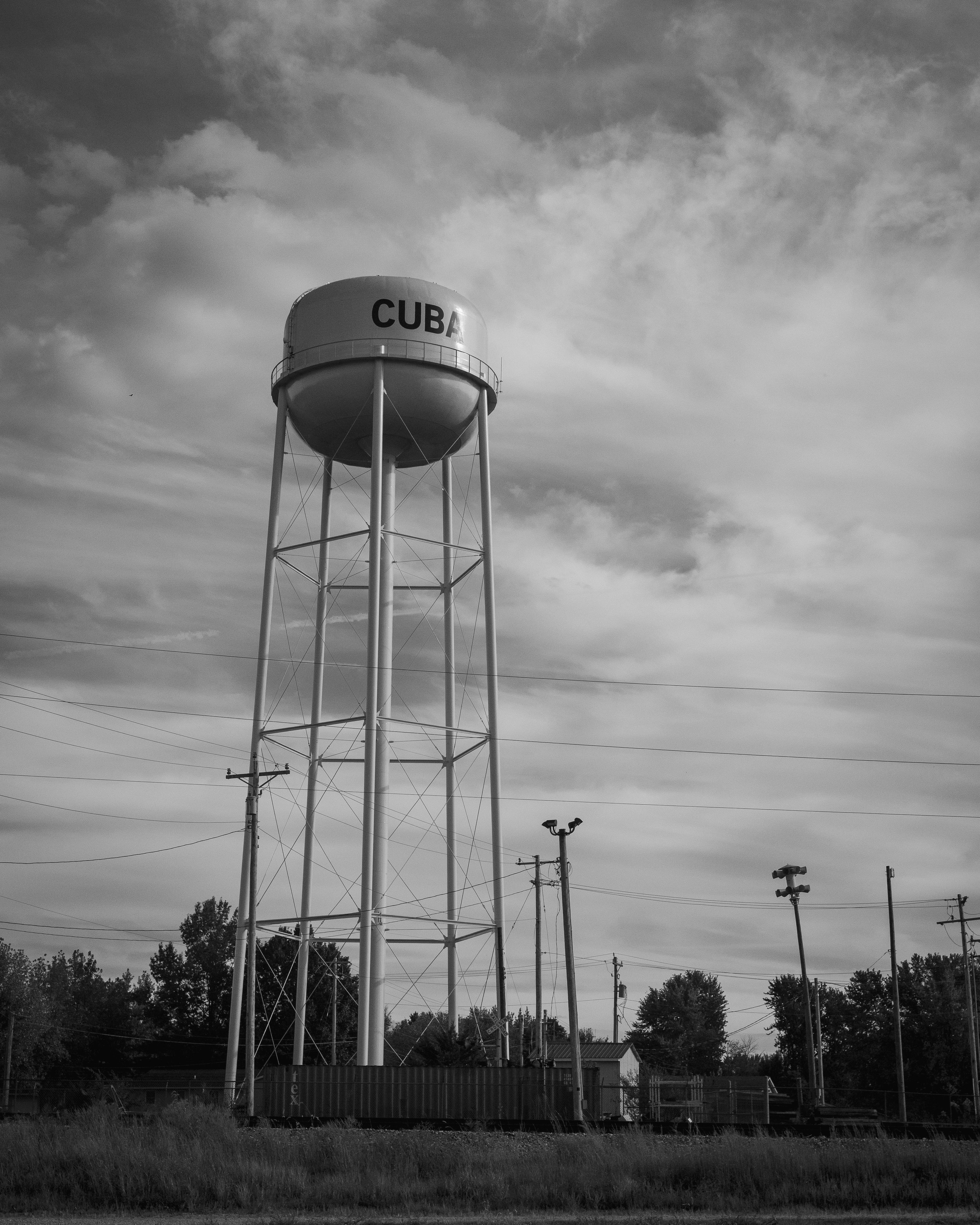 Un château d’eau portant l’inscription « CUBA », avec une structure métallique et des câbles, se dresse sur fond de ciel nuageux.