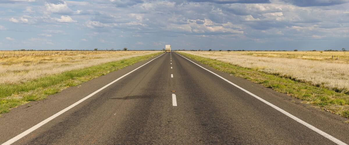 A long, straight road with a white line down the center, flanked by grassy fields under a partly cloudy sky.