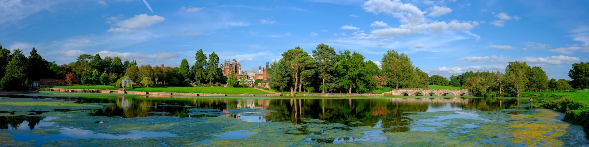 A serene lake with reflections, surrounded by lush greenery and a historic building in the background.