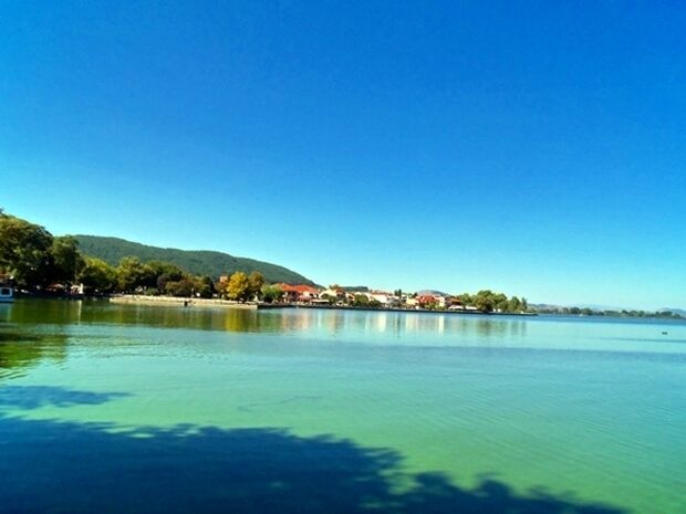 A calm lake with clear water, surrounded by trees and buildings on the shore.