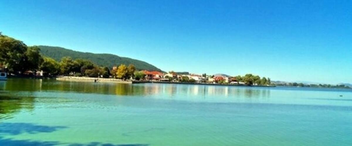 A calm lake with clear water, surrounded by trees and buildings on the shore.
