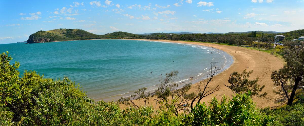 A coastal landscape with a sandy beach, clear turquoise waters, and green hills in the background.