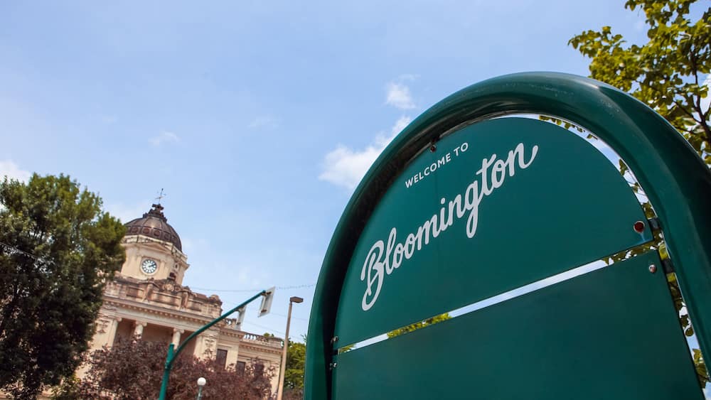 Un panneau vert sur lequel on peut lire « Bienvenue à Bloomington », une tour de l’horloge et des arbres en arrière-plan.