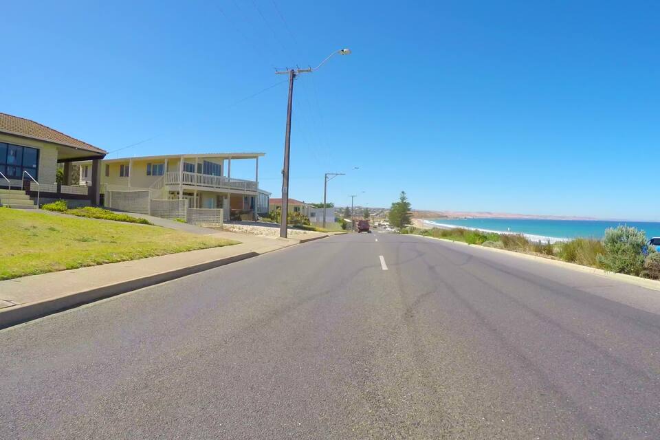 A coastal road with a clear view of the ocean and a residential area with houses and a car parked by the roadside.