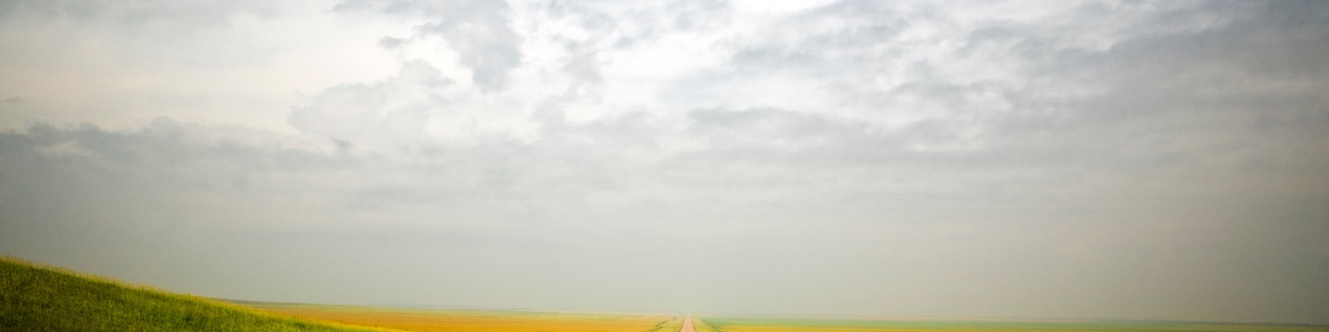 A long, straight dirt road flanked by green fields under a partly cloudy sky.