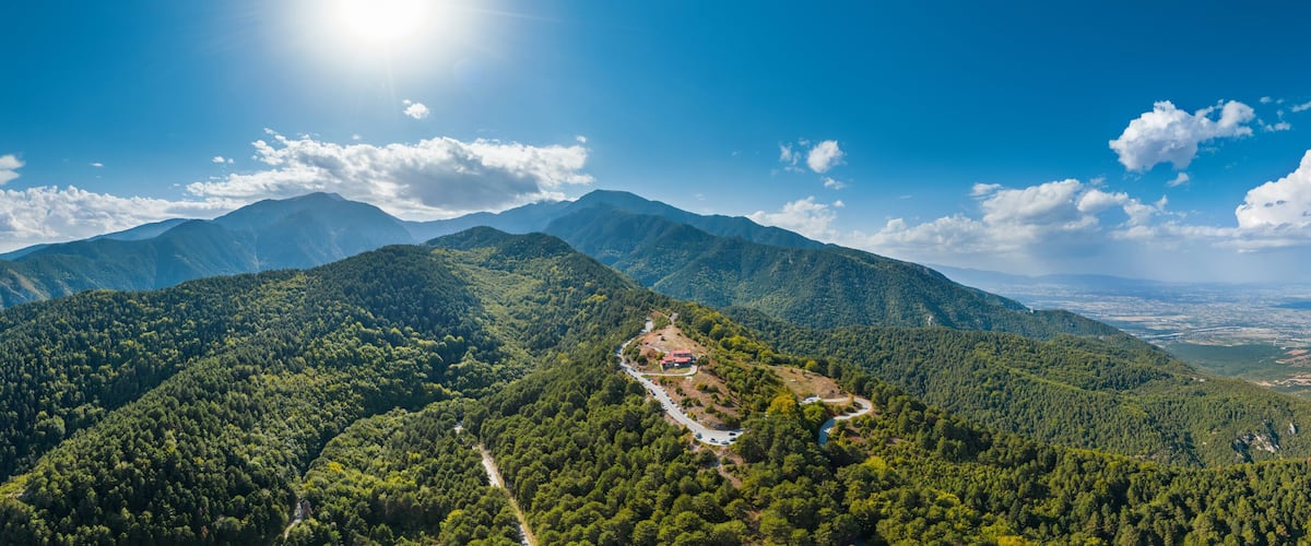 A mountainous landscape with dense forests, a winding road, and a distant cityscape.