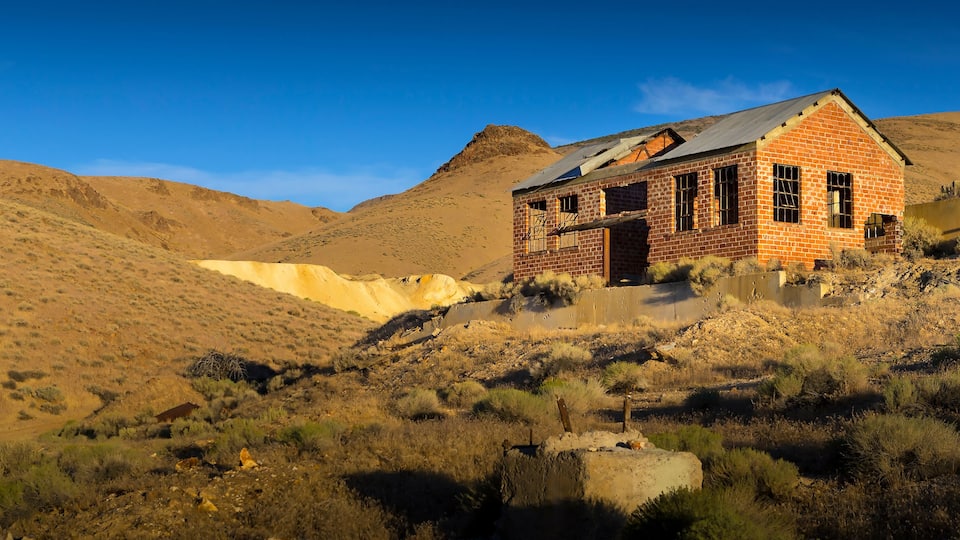 A dilapidated brick building on a hillside with sparse vegetation and a clear blue sky.