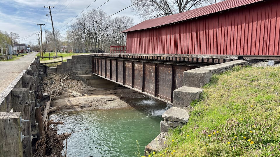 A red covered bridge over a narrow water channel, surrounded by grassy areas and trees.