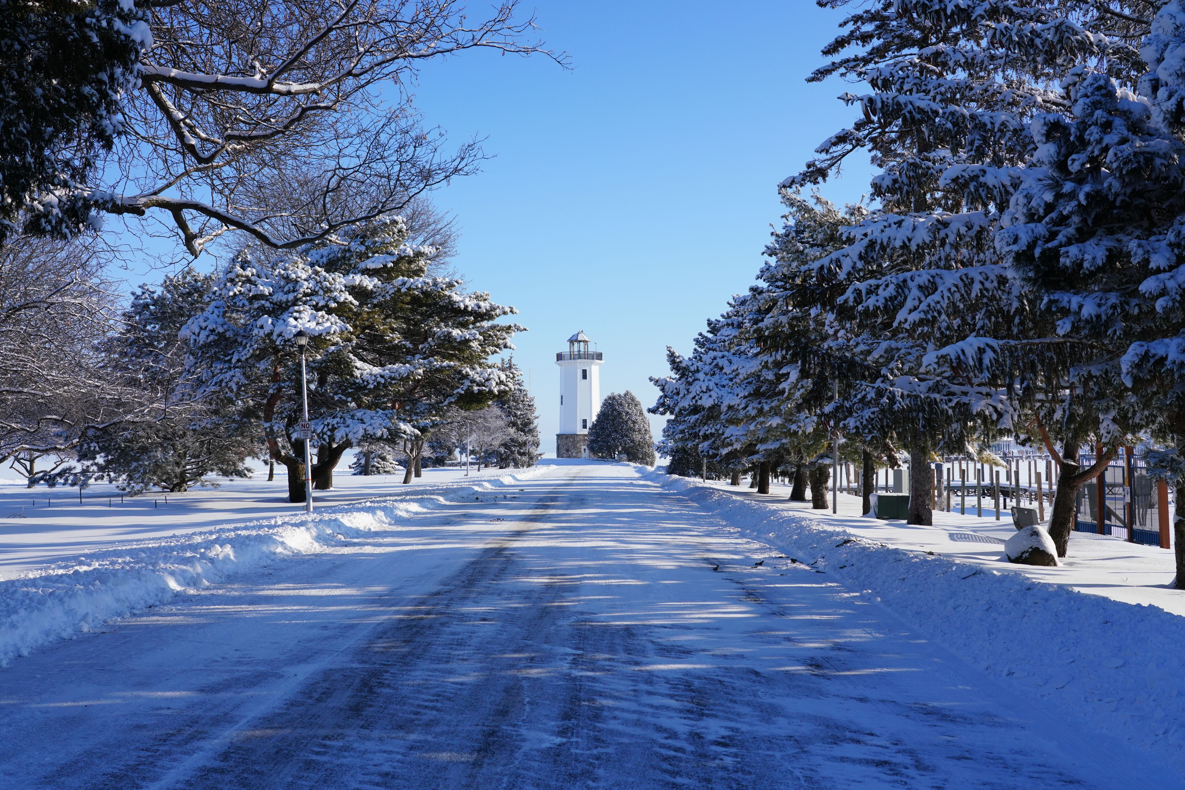 A snow-covered road with a lighthouse in the distance, surrounded by snow-laden trees.