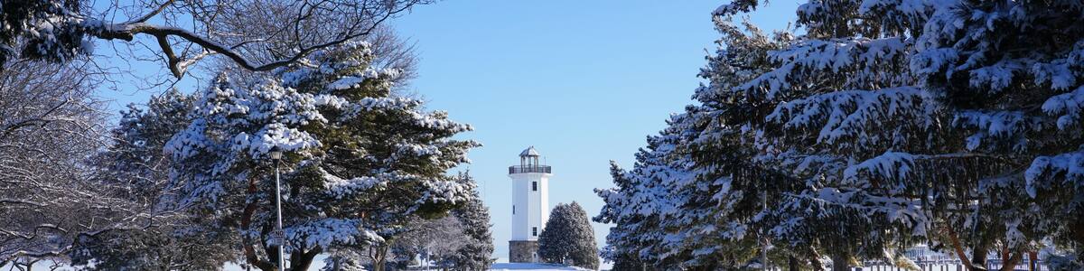 A snow-covered road with a lighthouse in the distance, surrounded by snow-laden trees.