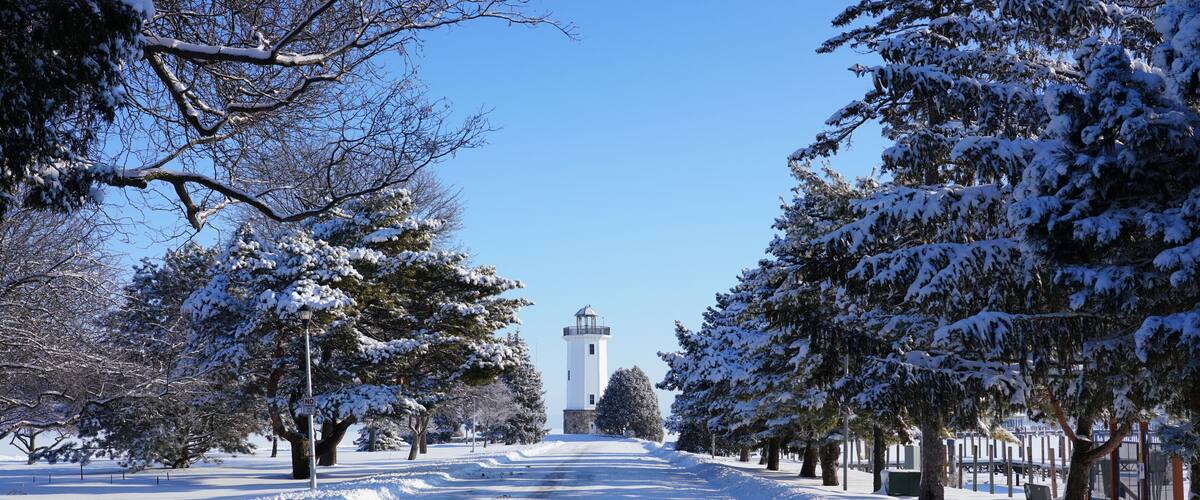 Una carretera cubierta de nieve con un faro a lo lejos, rodeada de árboles cargados de nieve.