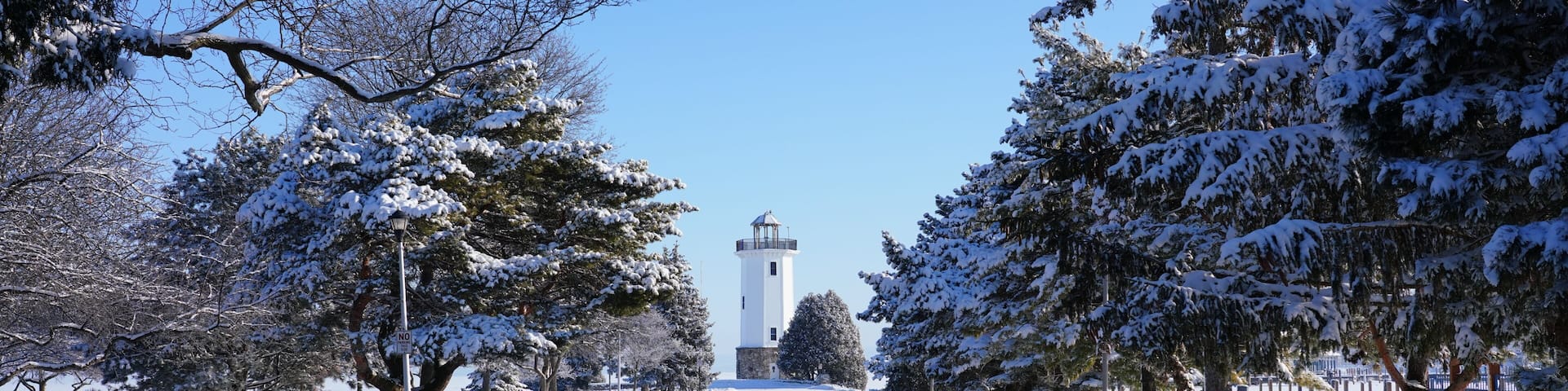 A snow-covered road with a lighthouse in the distance, surrounded by snow-laden trees.