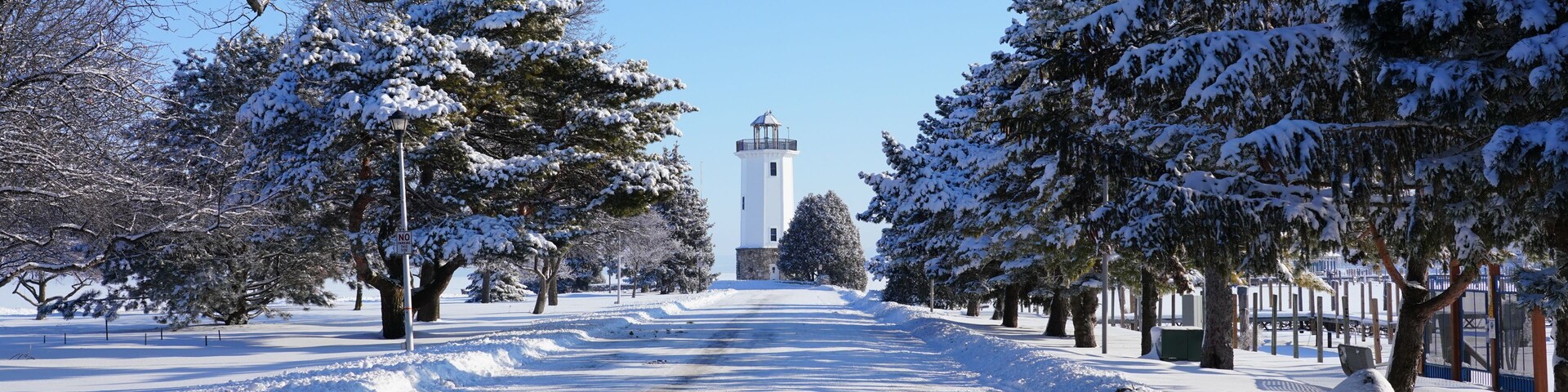 Uma estrada coberta de neve com um farol ao longe, cercada por árvores carregadas de neve.