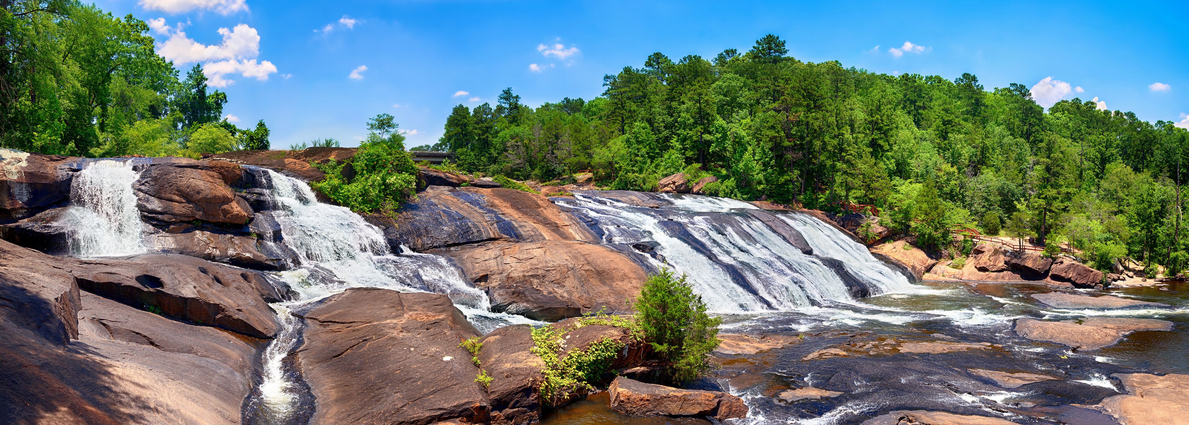 A multi-tiered waterfall cascading over large rocks, surrounded by dense green forest.