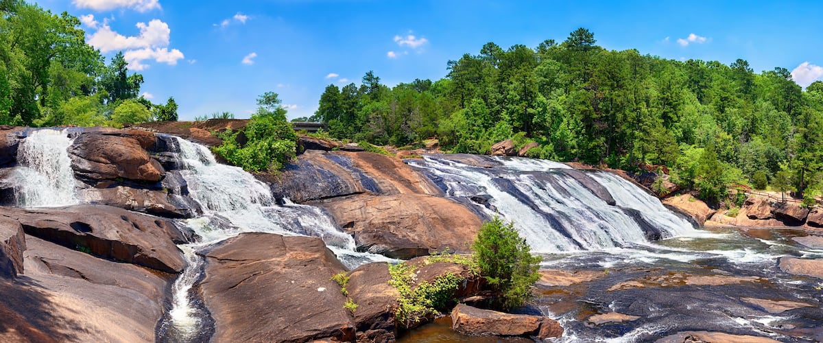 A multi-tiered waterfall cascading over large rocks, surrounded by dense green forest.