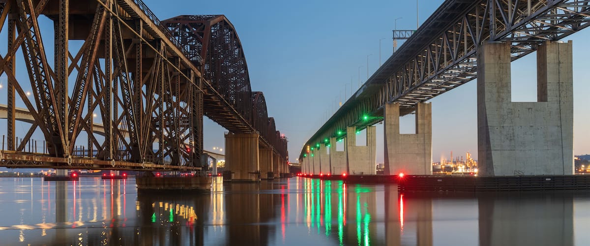 Un pont d’acier enjambant une étendue d’eau calme, où se reflètent les lumières, une structure en béton et un ciel dégagé.