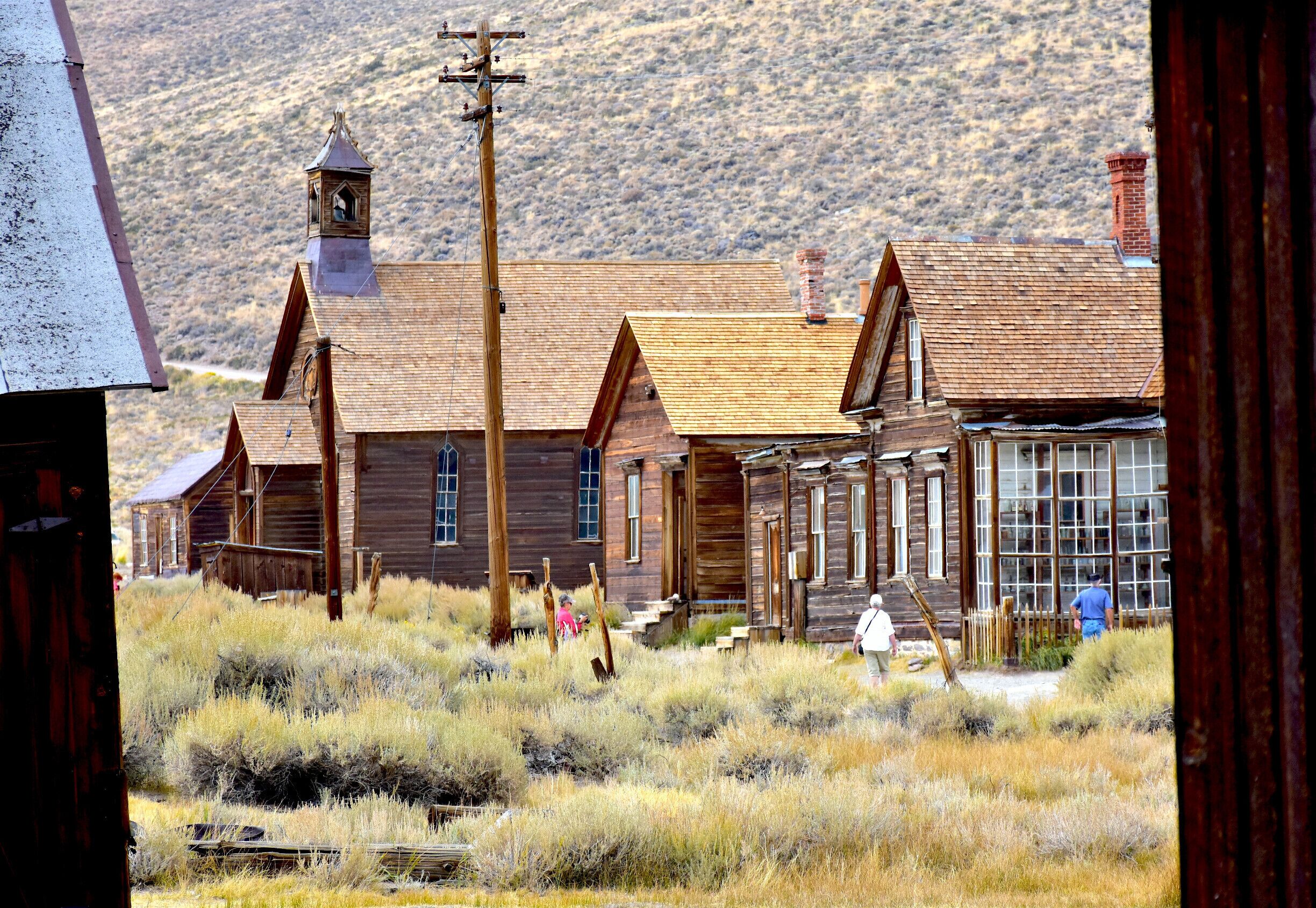A historic wooden building with a bell tower, surrounded by dry grassland and mountains in the background.