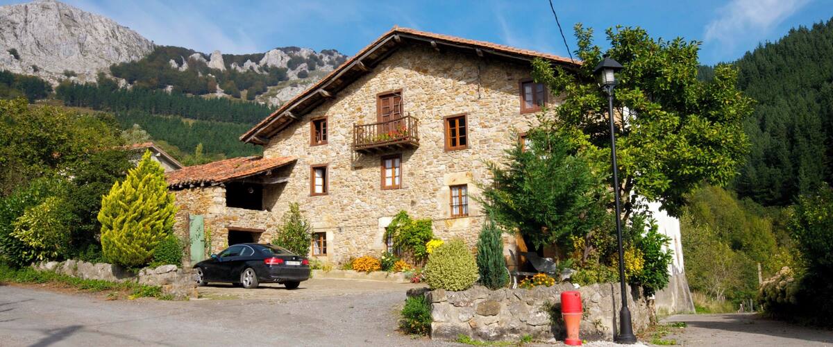 A stone house with a car parked in front, surrounded by trees and mountains.