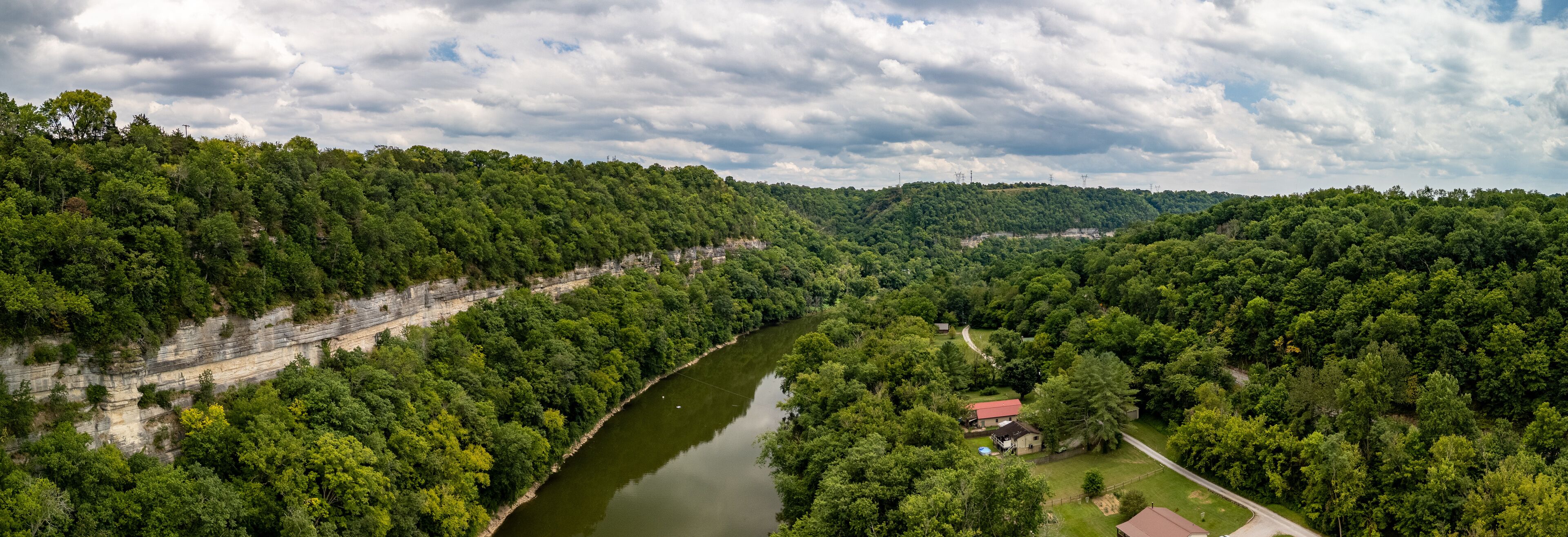 A river winding through a densely forested valley with a house and a red-roofed building near the water's edge.