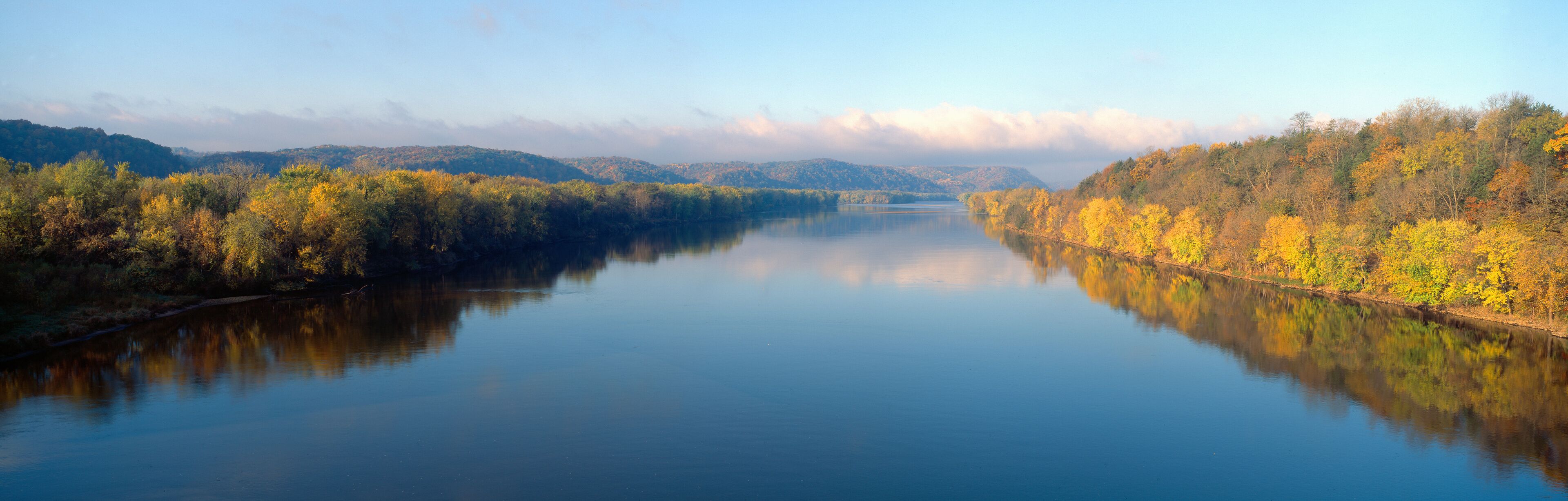 A calm river reflecting autumn foliage, surrounded by hills.