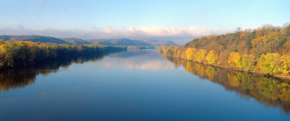 A calm river reflecting autumn foliage, surrounded by hills.