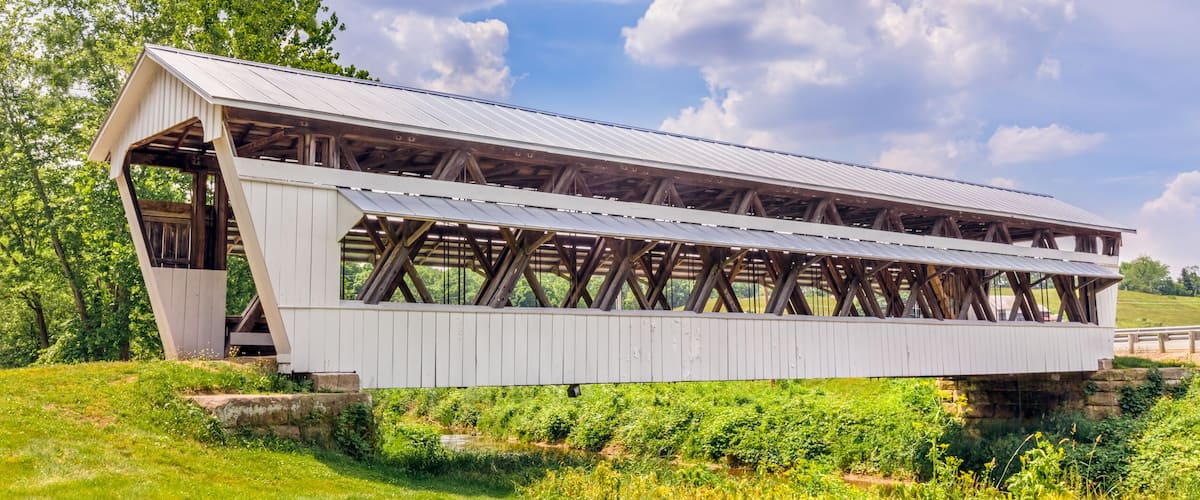 A covered bridge with a white exterior and wooden beams, spanning a grassy area with trees in the background.