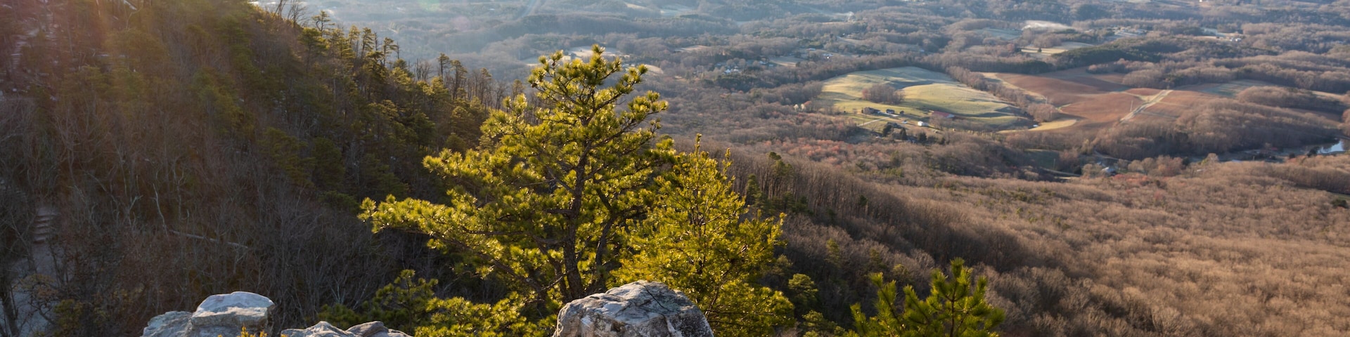 A mountainous landscape with a prominent rocky outcrop in the foreground and a valley with scattered trees in the background.