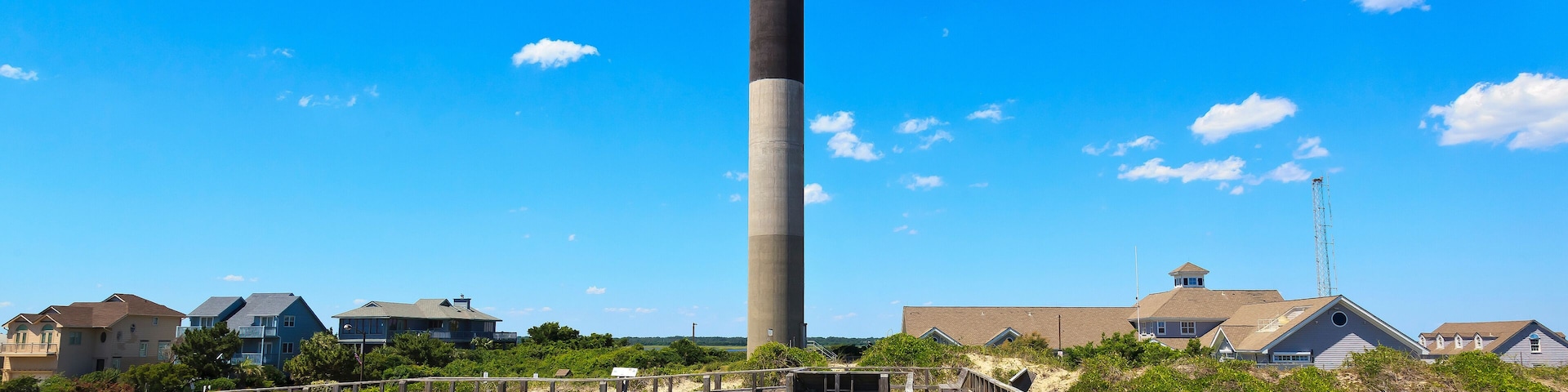 A wooden boardwalk leading to a tall chimney, surrounded by houses and greenery.