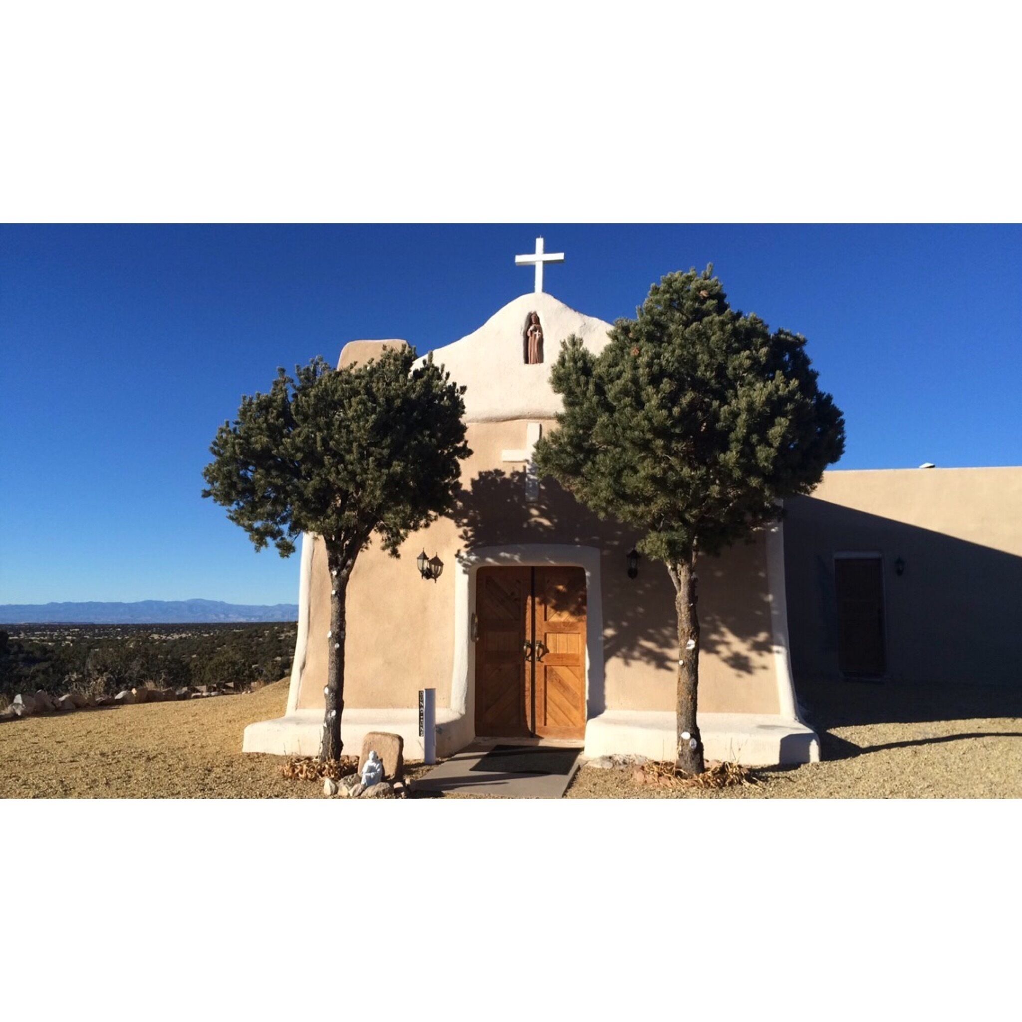 A small church with a cross on top, two trees in front, and a clear blue sky.