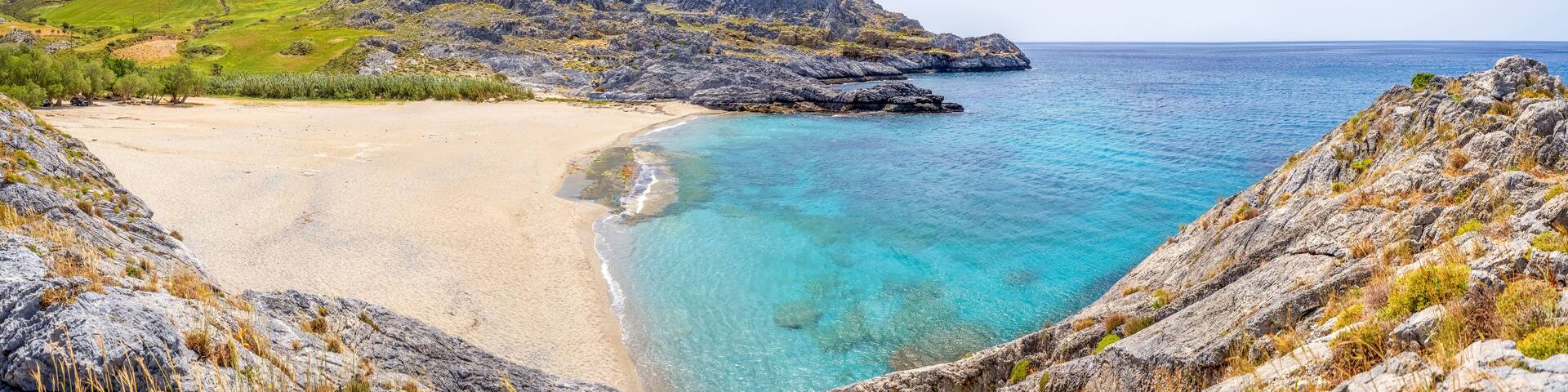 A coastal landscape with a sandy beach, clear turquoise waters, rocky cliffs, and a green hill in the background.