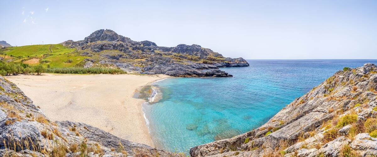 A coastal landscape with a sandy beach, clear turquoise waters, rocky cliffs, and a green hill in the background.
