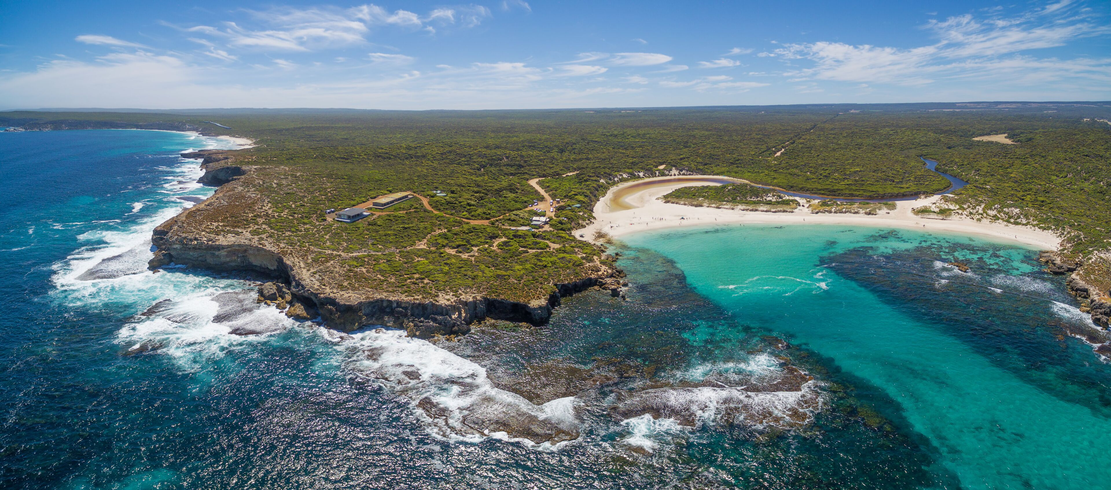 An aerial view of a coastal landscape with a beach, clear turquoise waters, and a dense forested area.
